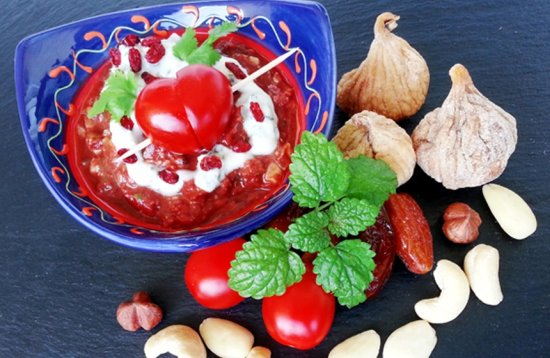 Bunte, dekorativ angerichtete Speise auf dunklem Untergrund: Eine blaue Schale mit Tomatensoße, darauf Cherry-Tomaten in Herzform. Arrangiert sind: getrocknete Feigen, kleine Tomaten, grüne Blätter, Nüsse und Knoblauch.