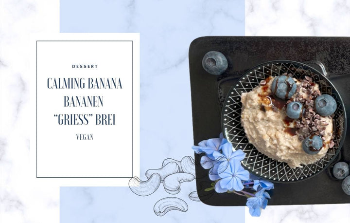 Foto eines veganen Desserts namens „Calming Banana Bananen ‘Griess’ Brei“ in einer schwarzen Schale, garniert mit Heidelbeeren, Kakaonibs und Schokoladensauce, auf einem schwarzen Tablett mit blauen Blumen daneben