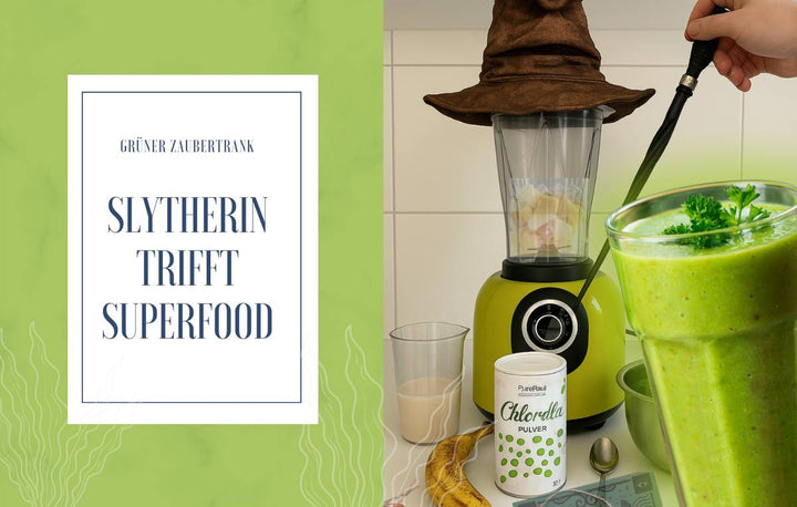 Ein grüner Smoothie steht neben einem Mixer mit Zauberhut und einer Dose Chlorella-Pulver. Die Grafik trägt den Schriftzug „Slytherin trifft Superfood“ und spielt mit einer magisch-grünen Optik.