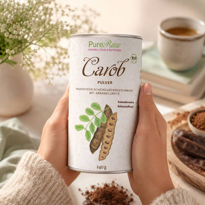 Zwei Hände halten eine Dose „Carob Pulver“ in einer gemütlichen Küchenatmosphäre mit einer Tasse im Hintergrund.